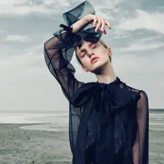 Modelka w czarnej sukience z tiulowymi rękawami, z kokardą na szyi, pozuje na tle pustkowia i nieba.