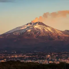 Etna, najwyższy wulkan Europy, dymi na tle zachodzącego słońca. U jego stóp rozciąga się miasto.