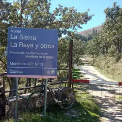 Señal de "Puerto de la Morcuera" junto a una bicicleta de montaña. Camino rural y árboles en un día soleado.