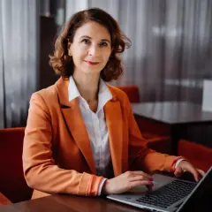 Kobieta w pomarańczowej marynarce pracuje na laptopie w eleganckim wnętrzu.