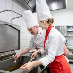 Deux chefs en cuisine, un homme et une femme, travaillent ensemble. L'homme, lunettes, semble superviser la jeune femme, qui porte un tablier rouge. Une scène parfaite pour une lettre de motivation second de cuisine.