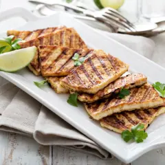 Grillowany ser halloumi, idealny jako marynata na grilla, podany z ćwiartkami limonki i natką pietruszki na białej tacy.