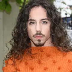 Michał Szpak w pomarańczowej bluzce z falbanami, z burzą loków i charakterystyczną bródką. Jego wiek nie wpływa na jego styl.