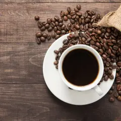 Aromatic coffee beans spill from a burlap sack onto a wooden table, next to a white cup filled with dark coffee. Poczuj kofeina działanie!