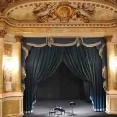 Złote zdobienia i bogate zasłony tworzą majestatyczne oświetlenie sceny teatralnej. Na scenie dwa stołki i pulpit nutowy czekają na artystów.