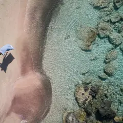 En person med en blå handduk på en rosa strand på Kreta. Klart turkost vatten med stenar.