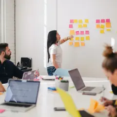 Zespół pracuje nad projektem, kobieta wskazuje na karteczki na tablicy. To może być zadanie 3 matura ustna angielski.