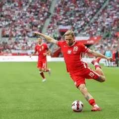 Piłkarz w czerwonej koszulce wykonuje piłkarskie dośrodkowanie, celując w pole karne. Tłum kibiców na trybunach.
