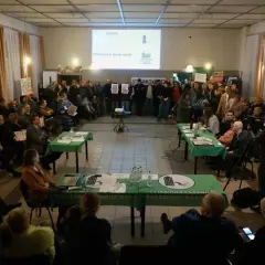 Ludzie siedzą i stoją na spotkaniu, na stole leżą dokumenty i laptop.
