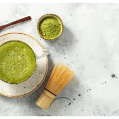 Jak pić herbatę matcha? Na stole leży spieniona zielona herbata w filiżance, obok bambusowa miotełka, łyżeczka z proszkiem i miseczka z matchą.