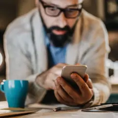 Mann mit Brille und Bart tippt auf Smartphone. Neben ihm eine blaue Tasse und ein Taschenrechner. Er sucht vielleicht, was ist die bankleitzahl.