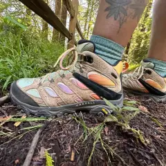 A person's feet wearing tan Merrell hiking boots with colorful accents and teal socks on a forest trail. These boots fit snugly, offering support for the hike.