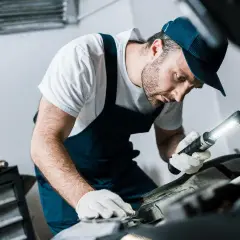 Mechanik w białej koszulce i granatowym kombinezonie, z latarką w ręku, sprawdza poziom płynu do sprzęgła w samochodzie.