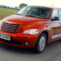 Pomarańczowy Chrysler PT Cruiser jedzie po drodze, za nim zielone pola i drzewa.