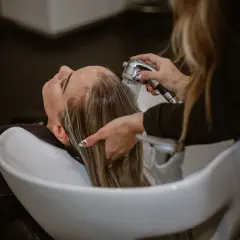 Una persona se lava el pelo con agua fría en un lavacabezas. El agua cae sobre su cabello rubio mientras una mano lo sujeta.