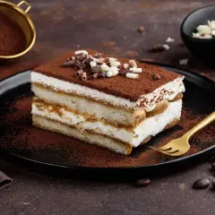 Una fetta di tiramisù con philadelphia, cosparso di cacao e riccioli di cioccolato, su un piatto nero con forchetta dorata.