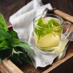 Aromatyczne picie pokrzywy w szklance, obok świeże liście na drewnianej tacy.