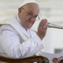 Papież Franciszek w białej szacie siedzi na krześle, trzymając mikrofon i kartkę.