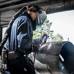 Spawacz wykonuje spawanie elektrodą w pionie na dużym metalowym elemencie. Widać iskry i łuk spawalniczy.