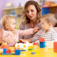Dzieci bawią się klockami przy stole, a opiekunka patrzy na nich z uśmiechem. Dofinansowanie do żłobka ułatwia rodzicom opiekę nad maluchami.