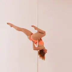 Kobieta w pomarańczowym stroju wykonuje skomplikowaną figurę na pionowym słupku, przypominającą flagę pole dance.