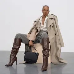 Eleganckie stylizacje z koszulą oversize: modelka w beżowym prochowcu, szarych spodniach, brązowych kozakach i czarnej torebce.