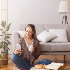 Uśmiechnięta kobieta z telefonem i kawą na podłodze, obok książka i rośliny. Czas na 5 pytań zdrowego myślenia.