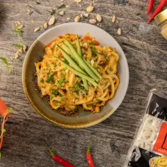 Pyszny makaron długi udon w kremowym sosie orzechowym, udekorowany ogórkiem i szczypiorkiem. Obok słoik tahini i paczka makaronu.