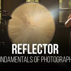 A large, round reflector is used to bounce light in this photography setup. A woman stands in the background, her hands raised.