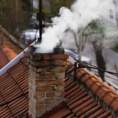 Rauch steigt aus einem alten Ziegelschornstein auf. Die Ziegel sind abgenutzt, was auf eine mögliche Schornsteinversottung durch Regen hindeutet.