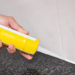 Mano aplicando silicona blanca con pistola en juntas de azulejo. Guía práctica de como aplicar silicona en juntas para un acabado perfecto.
