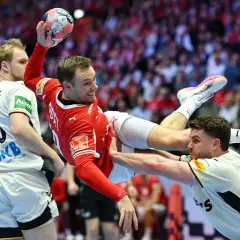 Piłkarz ręczny w czerwonej koszulce skacze do rzutu, broniony przez dwóch zawodników w białych strojach.