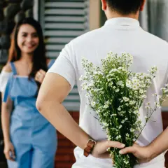 Mężczyzna z bukietem kwiatów za plecami, zbliżający się do uśmiechniętej kobiety. To piękny przykład pisania z innymi będąc w związku.