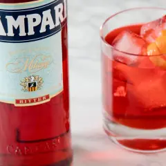 Butelka Campari i szklanka z czerwonym drinkiem, lodem i skórką pomarańczy. Idealny aperitif, który może być bazą dla drinków z Aperolem.