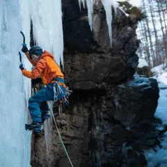 Wspinaczka górska po lodospadzie. Alpinista w pomarańczowej kurtce i niebieskich spodniach wspina się po zamarzniętej ścianie, używając czekana i raków.