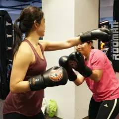 Two women spar in a boxing gym. One woman in a maroon tank top throws a punch, while the other in a pink shirt dodges. They both wear boxing gloves, ready for a cruiser weight bout.
