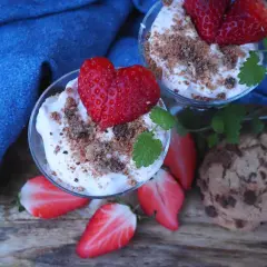 Två glas med jordgubbscheesecake, toppade med färska jordgubbar och smulor. En chokladkaka och jordgubbsbitar ligger bredvid.