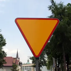 Żółty, trójkątny znak ustąp pierwszeństwa z czerwoną obwódką, stojący na tle drzew i nieba.