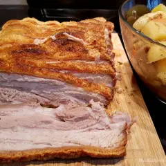 Ein butterzarter Schweinebauch mit knuspriger Kruste liegt auf einem Holzbrett, daneben eine Schüssel mit Bratkartoffeln und Gemüse.