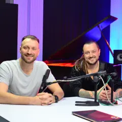 Alek Baron i jego kolega siedzą przy stole w studiu, gotowi do nagrania.