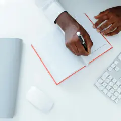 Un homme écrit une lettre réclamation frais bancaires dans un carnet. Un clavier et une souris sont à côté.