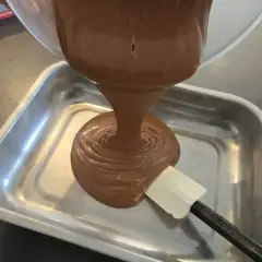 Une ganache au chocolat, inspirée par le Toblerone, est versée dans un plat métallique. Une spatule aide à l'étaler.