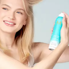 Mujer rubia sonriendo aplica champú seco para lavar pelo en seco, refrescando su melena.