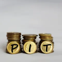 Trzy stosy monet, na których widnieją litery P, I, T. Symbolizują one rozliczenie podatku PIT.