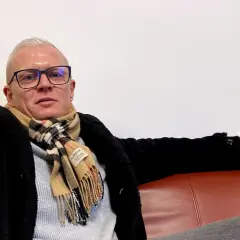 Robinson Alexander, med glasögon och en rutig halsduk, sitter avslappnat på en röd soffa.