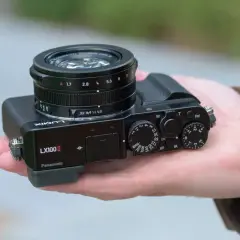A hand holds a Panasonic Lumix DMC-LX100K camera, showcasing its compact design and prominent lens.