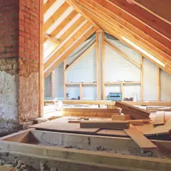 Drewniana konstrukcja dachu i komina, przygotowywana na poddasze użytkowe. Widać materiały budowlane i prowizoryczne półki.