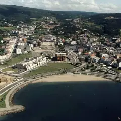 Vista aérea de la playa de Cee, con el mar azul, la arena dorada y la ciudad extendiéndose hacia las colinas verdes.