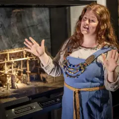 En kvinna i vikingakläder berättar vid en modell av en vikingagård på vikingamuseet.