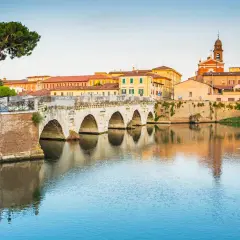 Il Ponte di Tiberio a Rimini, con i suoi archi in pietra che si specchiano sull'acqua calma, fa da sfondo a un paesaggio urbano colorato.